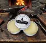 Two tins of Blacksmith Beard Balm on a wooden surface with blacksmith tools and a fireplace in the background.