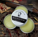 Three tins of Boomstick Beard Balm on a rustic wooden surface with a gun and other items in the background.