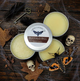 Buffalo Bill beard balm tins on a wooden surface with decorative elements like a deaths head moth and skull.