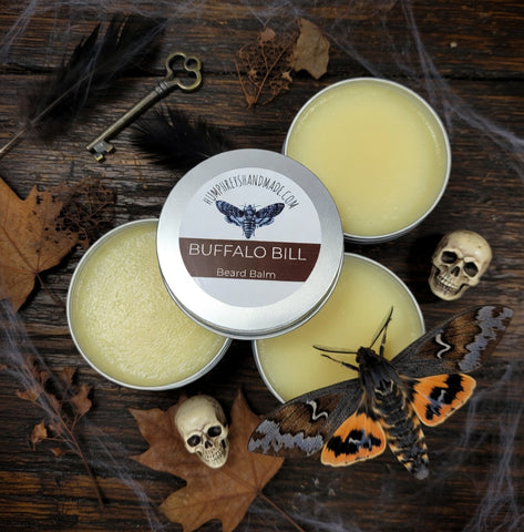 Buffalo Bill beard balm tins on a wooden surface with decorative elements like a deaths head moth and skull.