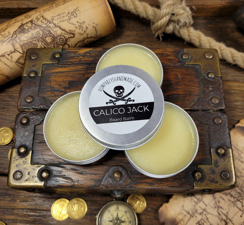 Three tins of Calico Jack beard balm on a wooden chest with coins and a map in the background.