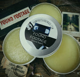 Three tins of 'Found Footage' beard balm on a rustic background with a 'Found Footage' movie poster.