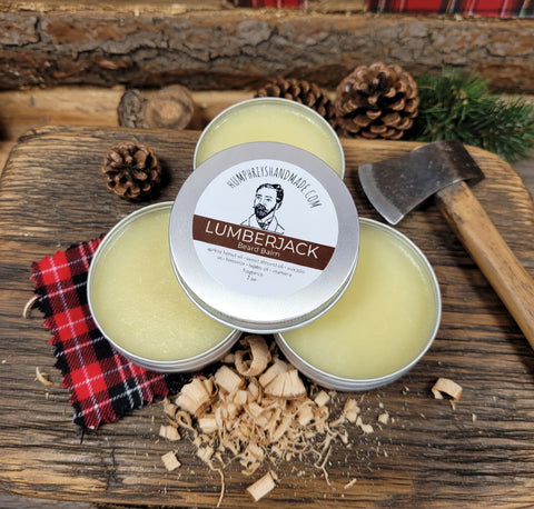 Three tins of Lumberjack beard balm on a wooden surface with a axe, plaid cloth, and pine cones.
